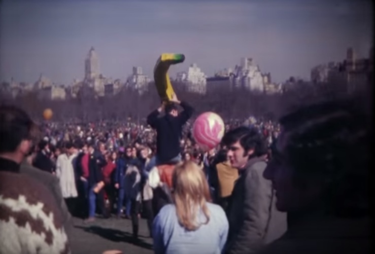 a huge crowd of people in Central Park, New York. One man sitting on the shoulders of another holds a huge model banana aloft