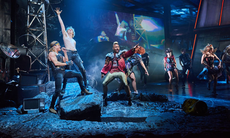 Scene from Bat Out Of Hell The Musical. Left to right - Giovanni Spano as Ledoux in profile with a mic, Andrew Polec as Strat - right open hand stretched skyward, Dom Hartley-Harris as Jagwire stridently pointing forwards, then several ensemble cast members - The Lost - in the background