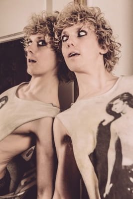 Andrew Polec as Strat in his Jim Steinman shirt, poses leaning against a mirror