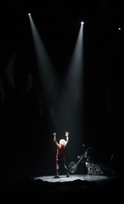 Scene from Bat Out Of Hell The Musical. Strat stands alone on stage, facing away, his arms are raised. The spotlights are on him and his motorcycle, all else is dark.