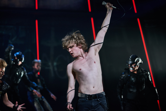 Scene from Bat Out Of Hell The Musical. Andrew Polec as Strat is in the foreground, bare chested and with microphone cord wrapped around both arms as he holds a microphone aloft while looking in the opposite direction. Behind him, members of The Lost and Falco's militia.