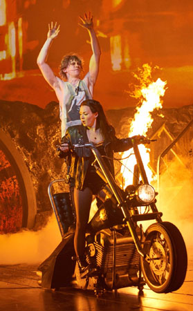Scene from Bat Out Of Hell The Musical. Strat and Raven are on a motorbike - Raven in front, Strat standing behind her, riding pillion. There is fire behind them, taller than a man, and the whole scene is tinted orange from the light of the flame