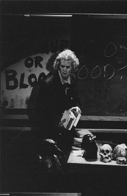 Barry Keating as The Historian. He holds several books. KETCHUP OR BLOOD? is scrawled on a blackboard behind him, a human skull and other objects on a desk beside him.